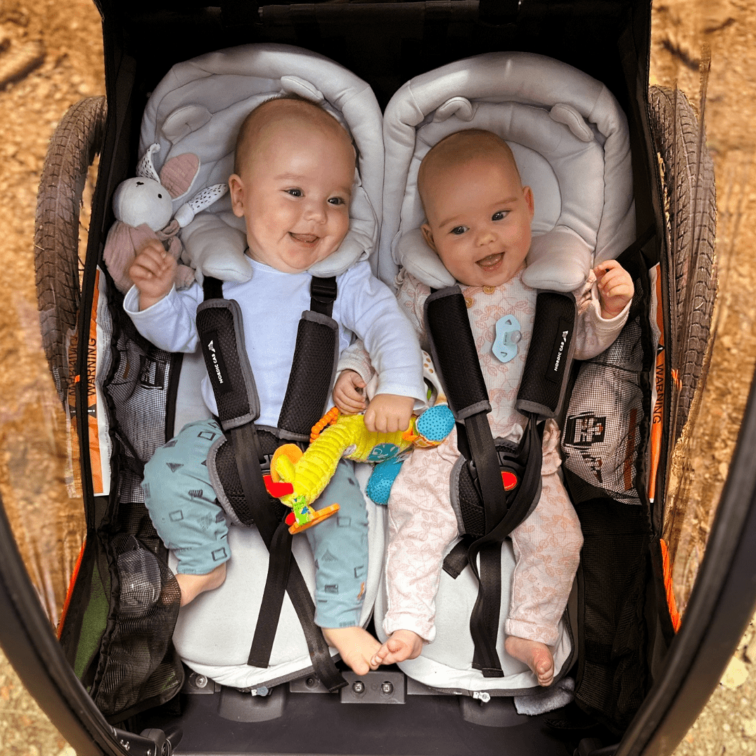 Two smiling babies in infant inserts inside a Nordic Cab bike trailer.
