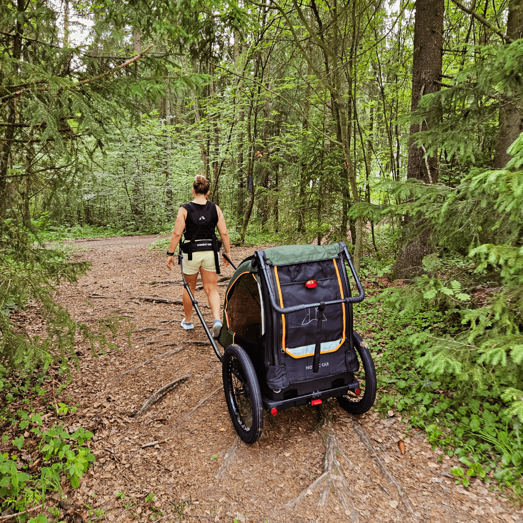 Woman pulling Nordic Cab bike trailer with harness along forest path.