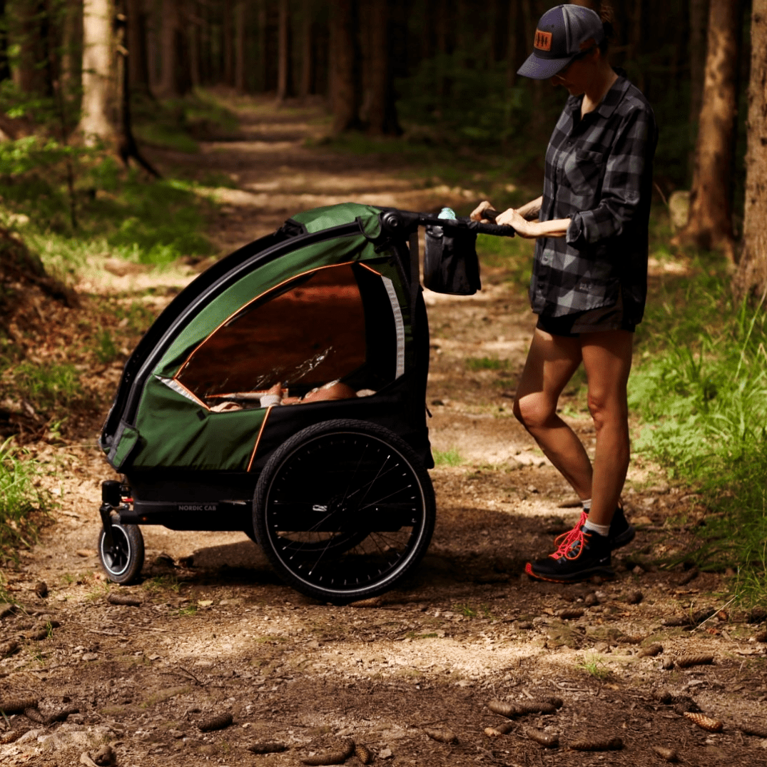 Woman pushing a Nordic Cab bike trailer in stroller mode on a forest trail.