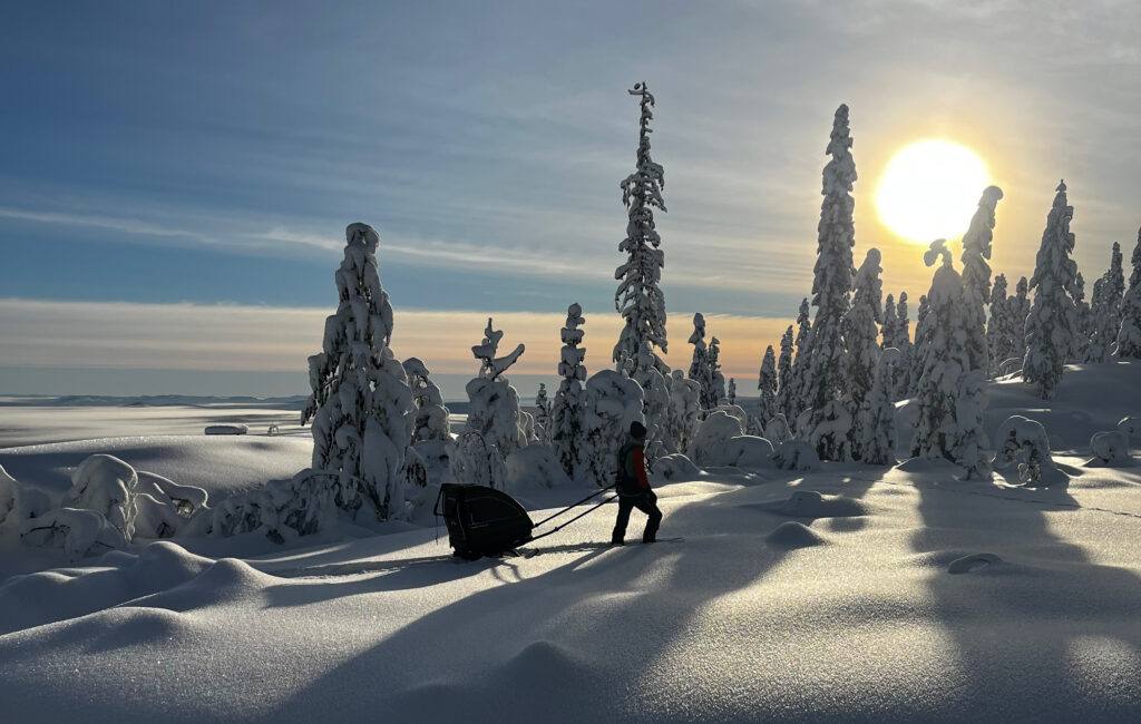 A person snowshoeing through deep, soft snow in Trysil, Norway, pulling a Nordic Cab sled in a snowy landscape under a bright winter sun